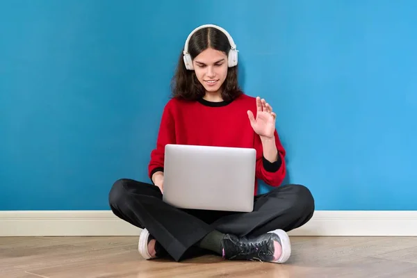 Teenage boy student in headphones using laptop for conference video call, sitting on floor on blue background. — Photo