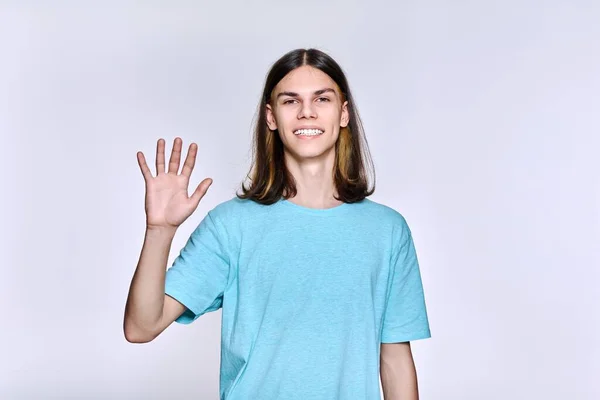 Positive teenage guy waving hand, looking at camera, light studio background —  Fotos de Stock