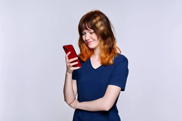Smiling teenage female looking at smartphone screen, on light studio background — Foto de Stock