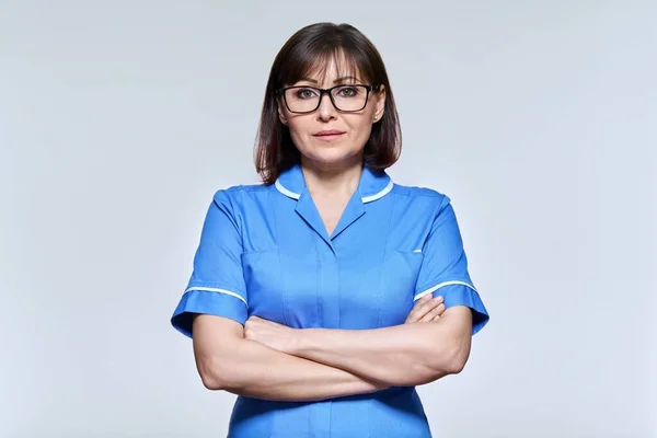 Portrait of middle aged nurse in blue uniform looking at camera on light studio background — Stock Photo, Image