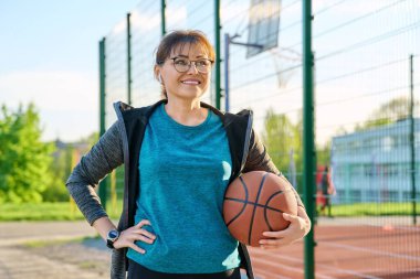 Orta yaşlı, elinde top olan aktif bir kadın, açık hava basketbol sahasının yanında.
