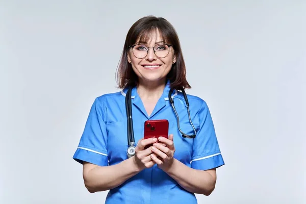 Portrait of female nurse with smartphone in hands, looking at camera on light background — Fotografia de Stock