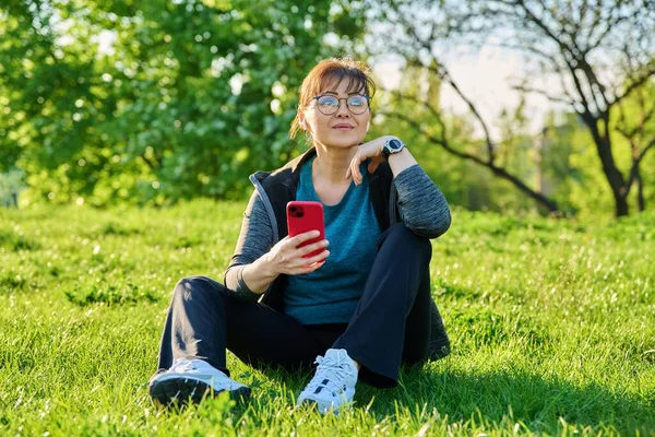 Gammal kvinna i hörlurar med smartphone lyssna musik, sitter på gräs — Stockfoto