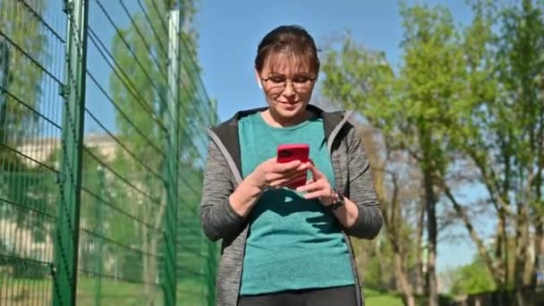 Mujer madura activa en ropa deportiva con teléfono inteligente, al aire libre — Vídeos de Stock