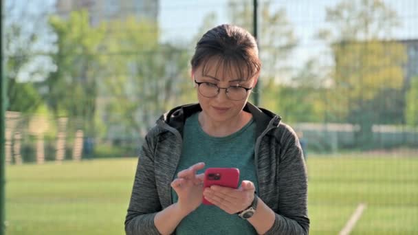 Mujer madura activa en auriculares de ropa deportiva con teléfono inteligente, al aire libre — Vídeos de Stock