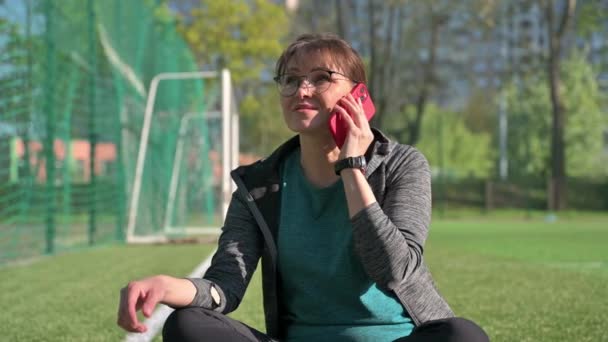 Middle aged woman in sportswear talking on the phone while sitting at sports outdoor stadium — Vídeos de Stock