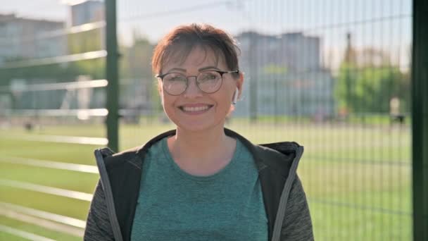 Glücklich lächelnde Frau mittleren Alters in Sportbekleidung blickt in die Kamera, Stadion-Hintergrund im Freien. — Stockvideo
