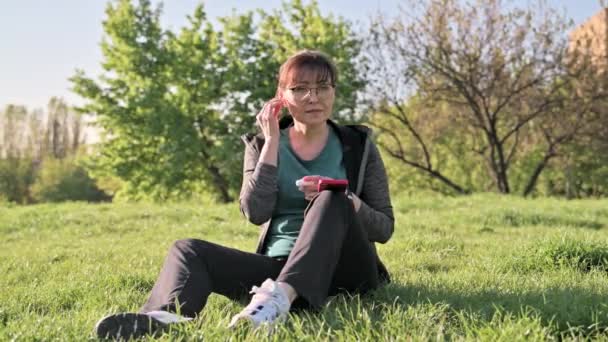 坐在草地上，头戴智能手机，听音乐的成熟女性 — 图库视频影像