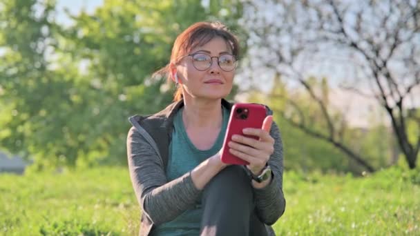 Ώριμη γυναίκα σε ακουστικά με μουσική ακρόασης smartphone, κάθεται στο γρασίδι — Αρχείο Βίντεο