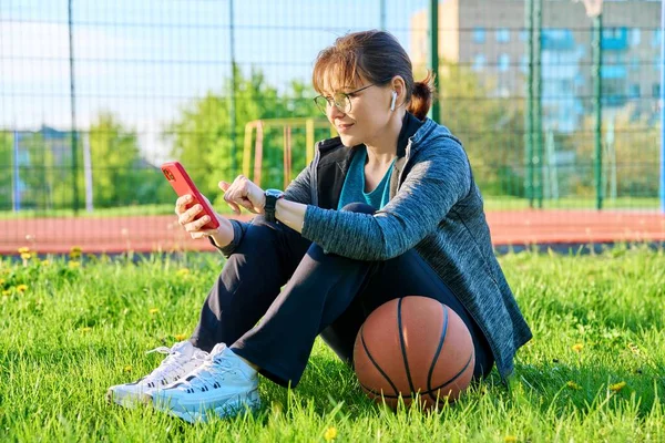 Ενεργή ώριμη γυναίκα με μπάλα μπάσκετ και smartphone, εξωτερική — Φωτογραφία Αρχείου