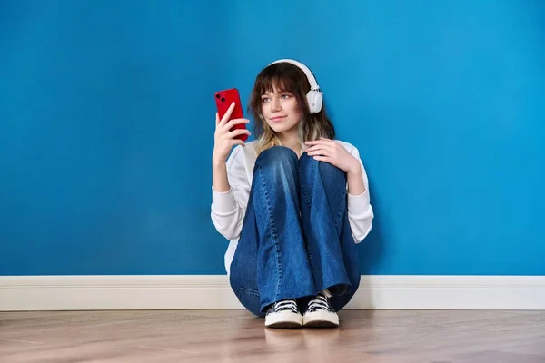 Підліткова жінка в навушниках зі смартфоном, що сидить на підлозі на синьому — стокове фото