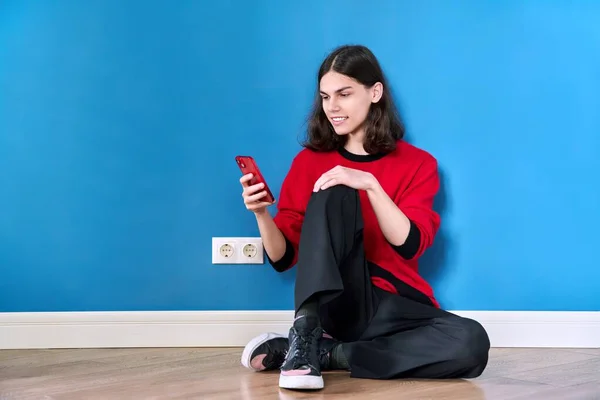 Teenage male with smartphone sitting on the floor on blue background — 스톡 사진