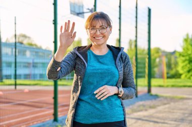 Pozitif orta yaşlı kadın elini sallayan kameraya bakıyor..