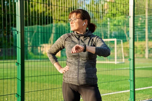 Spor giyimli aktif orta yaşlı bir kadın kol saatine bakıyor, egzersiz yapıyor.
