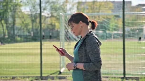 Aktív érett nő sportruházatban okostelefonnal, kültéri, profil nézet — Stock videók