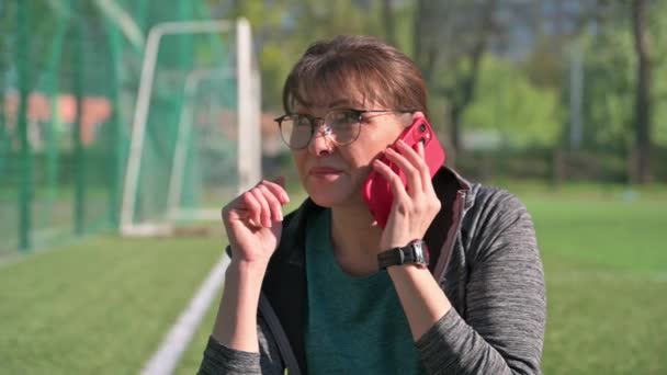 Kobieta w średnim wieku w odzieży sportowej rozmawia przez telefon siedząc na stadionie sportowym — Wideo stockowe