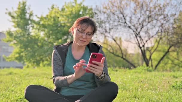 Reife Frau im Kopfhörer mit Smartphone Musik hören, sitzt auf Gras — Stockvideo