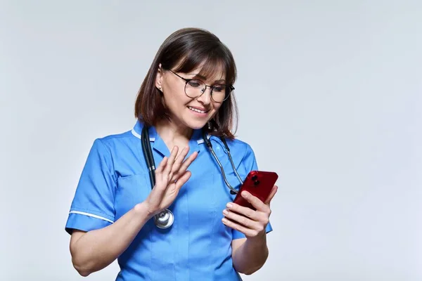 Enfermeira amigável acenando mão na tela do smartphone, no fundo do estúdio de luz — Fotografia de Stock
