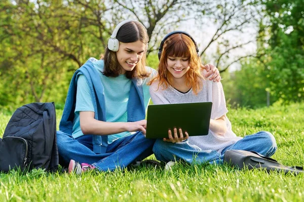 Два друга студенты, парень и девушка в наушниках, смотрят на ноутбук, сидят на траве — стоковое фото