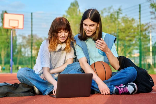 Φίλοι μαθητές που κάθονται στο γήπεδο μπάσκετ του σχολείου κοιτάζοντας φορητό υπολογιστή — Φωτογραφία Αρχείου