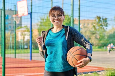 Orta yaşlı, elinde top olan aktif bir kadın, açık hava basketbol sahasının yanında.