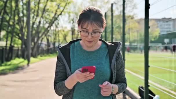Active mature woman in sportswear with smartphone, outdoor — Video