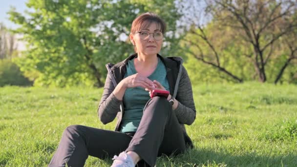 Reife Frau im Kopfhörer mit Smartphone Musik hören, sitzt auf Gras — Stockvideo