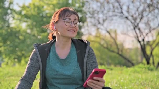 Mature female in headphones with smartphone listening music, sitting on grass — Vídeos de Stock