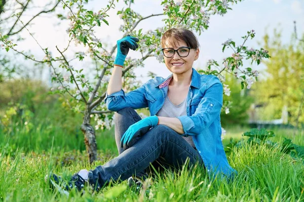 Positive beautiful mature female gardener sitting on the grass with blossoming apple tree. — Zdjęcie stockowe