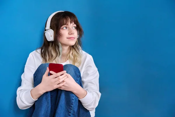 Teenage female in headphones with smartphone sitting on blue — Photo