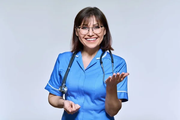 Позитивна усміхнена медсестра в синій формі дивиться на камеру на світлому фоні студії — стокове фото