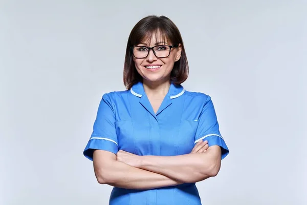 Portrait of middle aged nurse in blue uniform looking at camera on light studio background — Fotografia de Stock