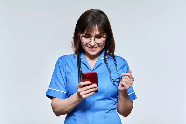 Retrato de enfermeira com smartphone nas mãos, olhando para o telefone no fundo claro — Fotografia de Stock