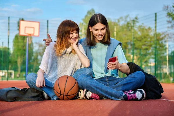 Student friends sitting on outdoor basketball court looking in smartphone — Stok fotoğraf