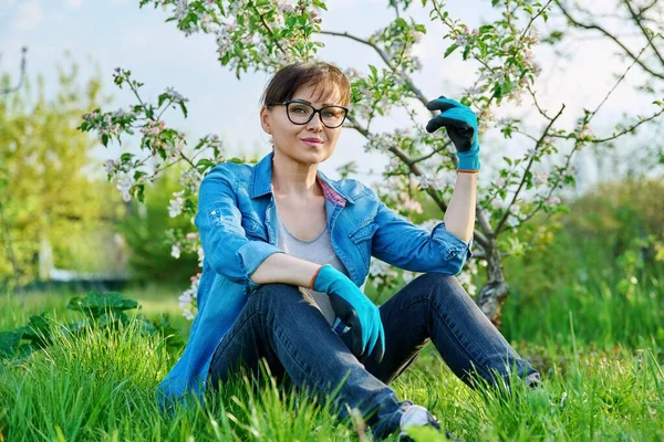 Positieve mooie volwassen vrouwelijke tuinman zittend op het gras met bloeiende appelboom. — Stockfoto