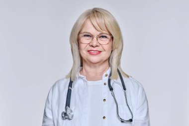 Steteskoplu kıdemli kadın doktor arka plandaki kameraya bakıyor.