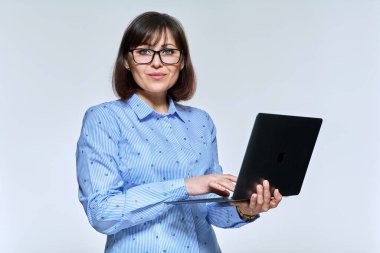 Orta yaşlı iş kadını dizüstü bilgisayar kullanıyor, arka plandaki kameraya bakıyor.