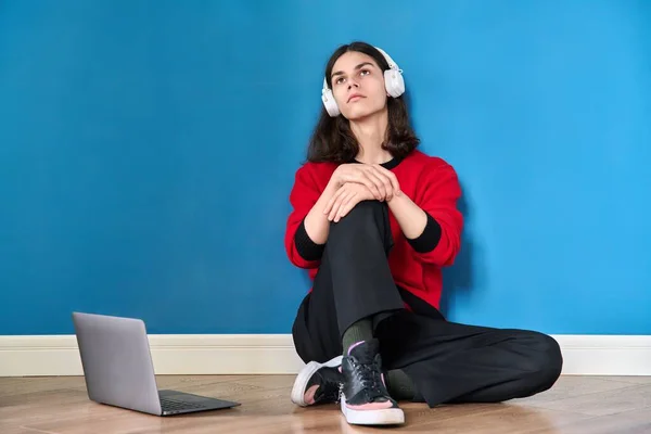 Jovem adolescente em fones de ouvido com laptop sentado no chão no fundo azul — Fotografia de Stock