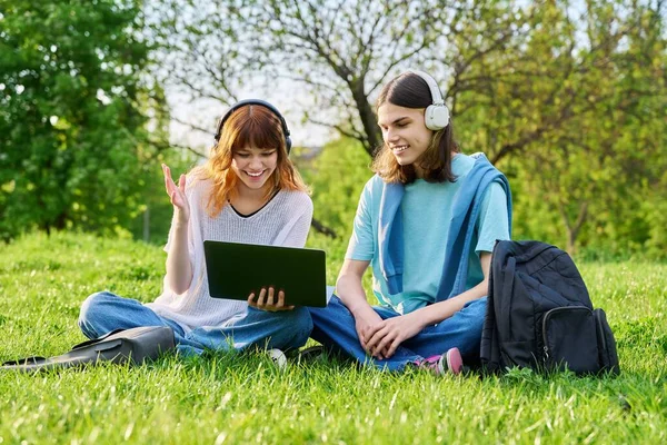 Два друга студенты, парень и девушка в наушниках, смотрят на ноутбук, сидят на траве — стоковое фото