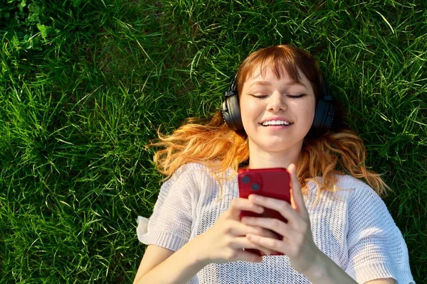 Top vista vermelho de cabelos jovens do sexo feminino em fones de ouvido com smartphone, na grama verde — Fotografia de Stock