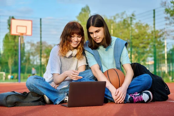 Φίλοι μαθητές που κάθονται στο γήπεδο μπάσκετ του σχολείου κοιτάζοντας φορητό υπολογιστή — Φωτογραφία Αρχείου