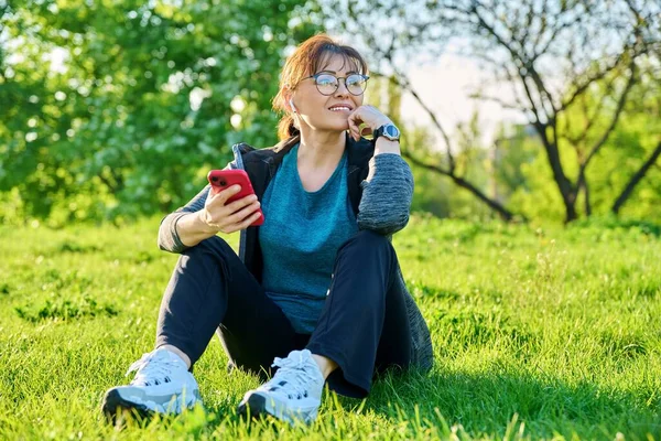 Mature female in headphones with smartphone listening music, sitting on grass — стоковое фото