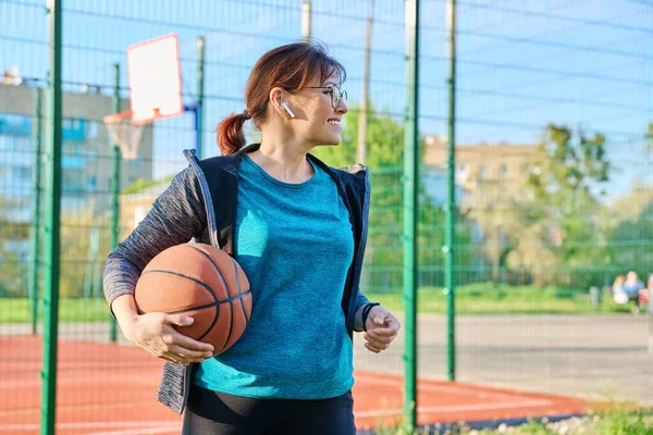 Середнього віку усміхнена активна жінка з м'ячем, біля відкритого баскетбольного майданчика — стокове фото