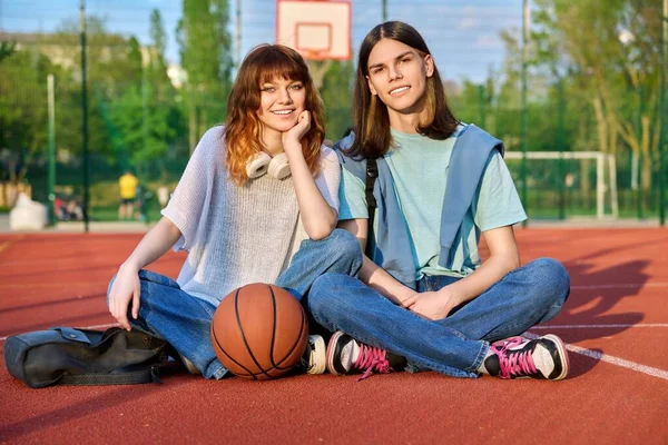 Barátok tini srác és lány néz kamera, ül a kosárlabdapályán — Stock Fotó