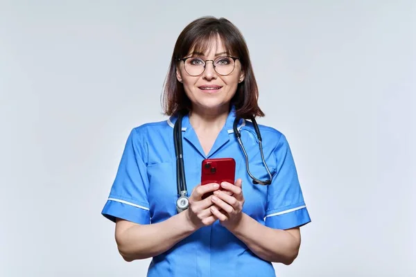 Portrait of female nurse with smartphone in hands, looking at camera on light background — Fotografia de Stock