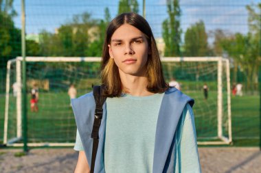Outdoor portrait of student boy 17, 18 years old in sunset on school football field stadium. Smiling positive male with backpack looking at camera. College high school, active sports healthy lifestyle