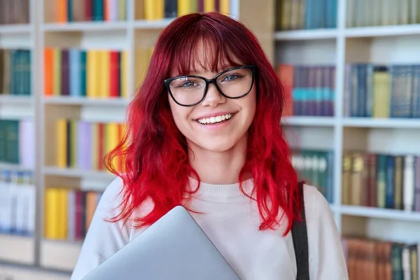 ノートパソコンのバックパックを持つ女性学生の肖像画 図書館でカメラを見て 眼鏡をかけ 歯で笑顔で赤い髪を染めて美しいファッショナブルな女の子 — ストック写真