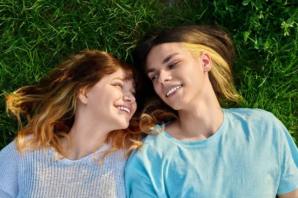 Couple of teenagers friends on green grass background. Happy guy and girl 17, 18 years old lying on lawn, top view. Youth, friendship, lifestyle, young people concept