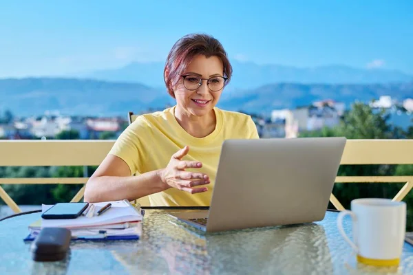 Middle aged woman in home office on outdoor terrace looking at laptop screen, talking on video call conference, online streaming. Remote work, virtual lesson, business, education, technology, people