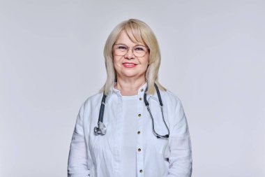 Steteskoplu gözlüklü gülümseyen yaşlı kadın doktorun portresi ışık stüdyo arka planında kameraya bakıyor. Meslek, tıp, sağlık hizmeti kavramı
